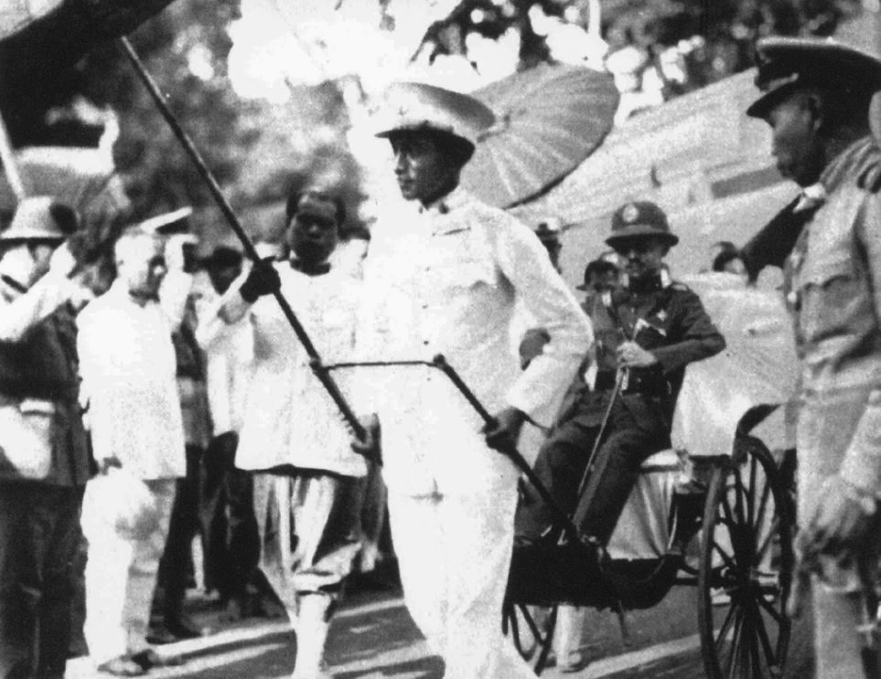 พระบาทสมเด็จพระปกเกล้าเจ้าอยู่หัว (ประทับรถลาก) ทอดพระเนตรร้านในงานฉลองรัฐธรรมนูญเมื่อวันที่ 12 ธันวาคม 2476 (ภาพจาก The Illustrated London News)